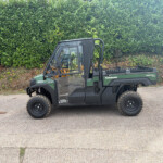 Side view of a green Kawasaki diesel 4x4 utility vehicle parked on a paved surface with a tall dense hedge in the background