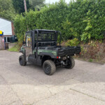 Green Kawasaki Diesel 4x4 utility vehicle in a parking area, with a yellow Russell’s sign storefront to the left and a tall green hedge behind it.