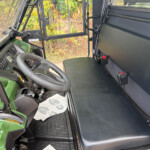 Interior of a green tractor cab showing steering wheel, dashboard controls, and black bench seat with a partition behind it, viewed from the left side near the windshield.