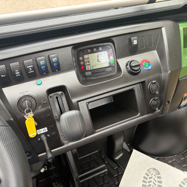 Truck dashboard with ignition key, digital display, and row of switches; green body panel visible outside.