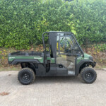 Green Kawasaki Diesel 4x4 utility vehicle parked on a paved surface in front of a dense green hedge; the open cargo bed and protective roll cage are visible over the left side of the vehicle.