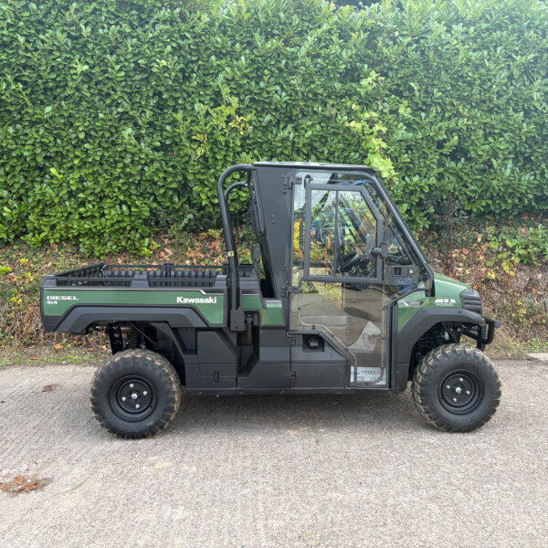 Green Kawasaki Diesel 4x4 utility vehicle parked on a paved surface in front of a dense green hedge; the open cargo bed and protective roll cage are visible over the left side of the vehicle.