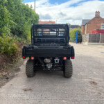 Rear view of a black Kawasaki off-road vehicle parked along a driveway beside a tall hedge on the left and houses on the right.