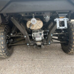 Rear view of an off-road vehicle showing the suspension, drivetrain, exhaust, and large knobby tires with red LED taillights visible on both sides.