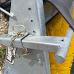 Close-up of a gray metal frame with a chain and padlock securing it, beside a large yellow bristle brush attachment.