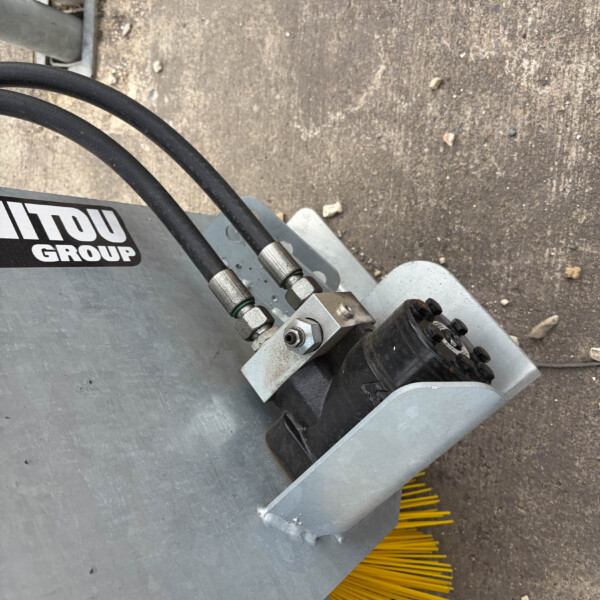 Close-up of an industrial machine with two black hydraulic hoses connected to a metal fitting, mounted on a gray metal chassis; yellow broom bristles are visible beneath.