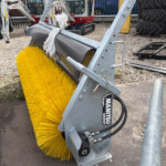 Close-up of a large yellow industrial street-sweeping brush mounted on a metal frame with hydraulic hoses, outdoors near excavators and stacked tires.