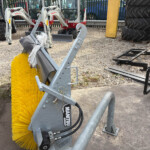 Close-up of a yellow circular street-sweeper brush attached to a gray metal frame, with tractors in the background on gravel.