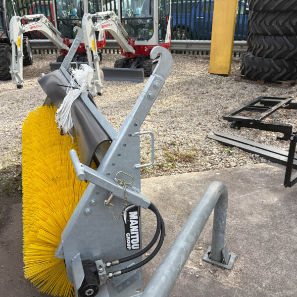Close-up of a yellow circular street-sweeper brush attached to a gray metal frame, with tractors in the background on gravel.