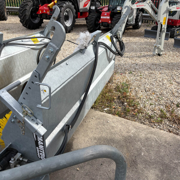 Heavy-duty construction attachment with hydraulic hoses and a yellow brush, mounted on a metal frame in a yard with tractors in the background.