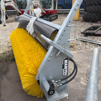 Close-up of a large yellow industrial street-sweeping brush mounted on a metal frame with hydraulic hoses, outdoors near excavators and stacked tires.
