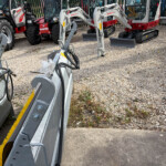 Parked construction machinery including excavator arms and red tractors on a gravel lot behind a fence.[]