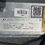 Close-up of a metal surface with a white barcode label and a QR code, beside a yellow safety warning label and a bolt head nearby.