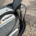 Close-up of hydraulic hose fittings and cables attached to a grey metal enclosure outdoors, with a gravel ground nearby.