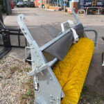 Industrial street-sweeper attachment with large yellow brush rollers mounted on a metal frame, outdoors in a lot.