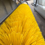 Close-up of bright yellow plastic bristles from a street-sweeper brush inside a metal housing.