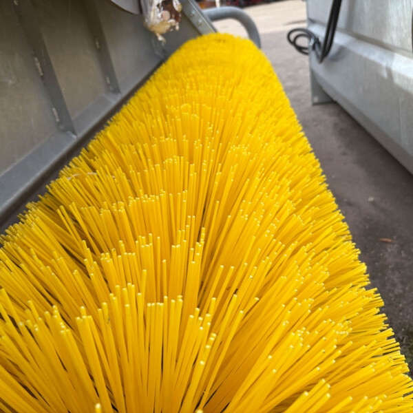 Close-up of bright yellow plastic bristles from a street-sweeper brush inside a metal housing.