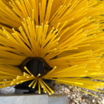Close-up of a yellow circular broom head with numerous stiff bristles radiating outward.