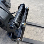 Close-up of a hydraulic cylinder and mounting pins on a heavy-duty forklift attachment at ground level.