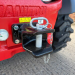 Close-up of a red tractor hitch with a silver lock and chain, warning sticker, and large tire in the background