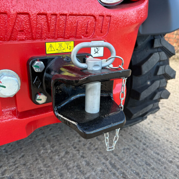 Close-up of a red tractor hitch with a silver lock and chain, warning sticker, and large tire in the background