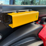 Close-up of a yellow rectangular control housing with two black knobs on top mounted to a dark machine chassis, part of heavy equipment.
