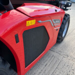 Close-up of a red Manitou front hood with a yellow safety warning sticker and large black radiator grille, tire nearby.