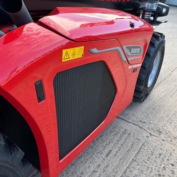Close-up of a red Manitou front hood with a yellow safety warning sticker and large black radiator grille, tire nearby.