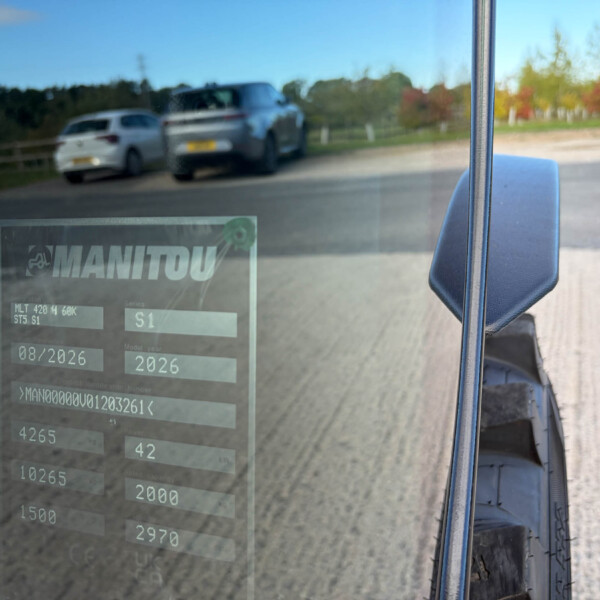 View from inside a vehicle looking through the windshield at a blurred road scene with a gray pickup and a white car parking along a fence; a translucent display shows the word MANITOU.