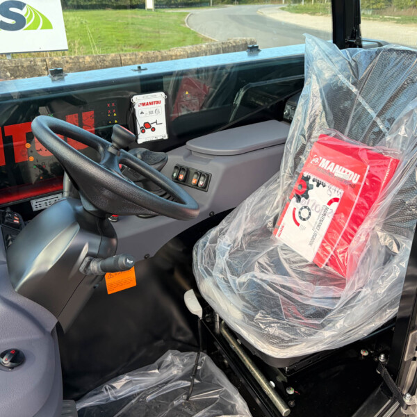 Cab interior of a construction machine; plastic-wrapped seat, steering wheel, and control panel visible with safety labels.