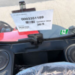 Vehicle dashboard with a white reference sheet taped to the windshield showing number 0003351189 and a barcode, plus a green emergency exit sticker on the right and two round speakers in the lower corners.