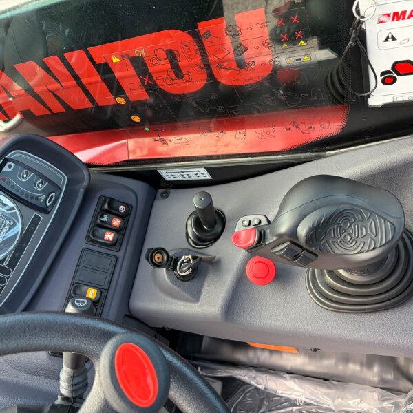 Controls and joystick in a heavy equipment cab with a digital display and red hazard markings visible outside the window (MANITOU).