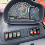 Dashboard of a vehicle showing a circular speedometer/tachometer cluster and rows of colored control buttons below.