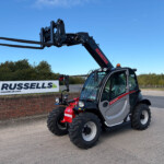 Telehandler with extended forks outdoors near a Russell's sign, showing a black machine and large tires under a clear blue sky.