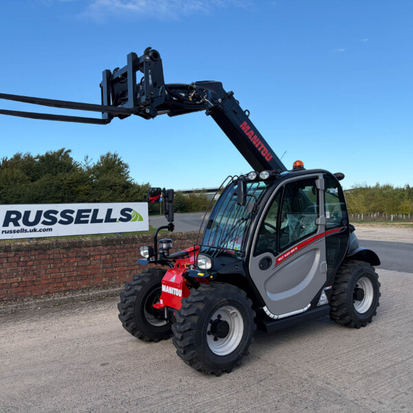 Telehandler with extended forks outdoors near a Russell's sign, showing a black machine and large tires under a clear blue sky.