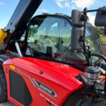 Close-up of a red Manitou MLT telehandler cabin with a large windshield, yellow lifting attachment, and hydraulic arms outdoors.