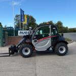 Compact utility vehicle with front forklift forks extended in a yard, blue tractors and flags in the background.