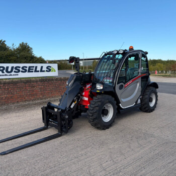 Compact forklift with extended forks parked on a paved area beside a brick wall and Russell's sign in the background.