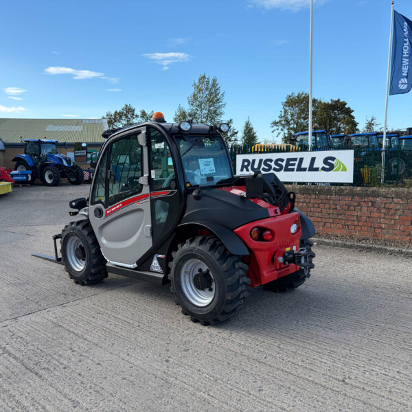 Red and black compact utility vehicle with an enclosed cabin in a dealership yard, with blue tractors and a Russell sign in the background.