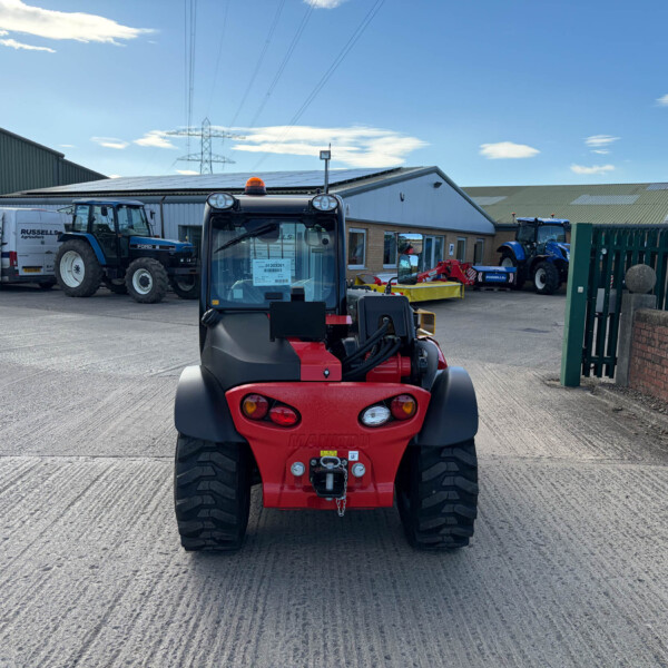 Front view of a red agricultural telehandler in the foreground, with tractors, a van, and farm buildings in the background under a blue sky.