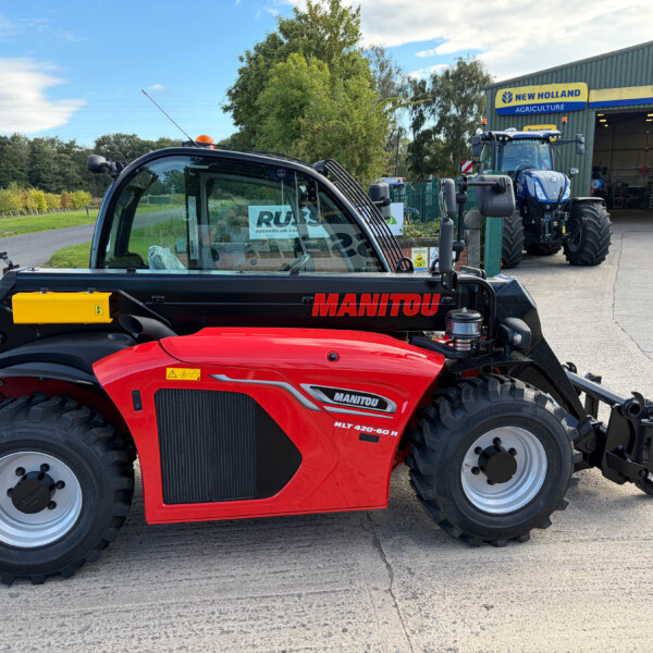 Red Manitou MLT 420-60 H telehandler parked in a farm equipment yard with a blue New Holland tractor inside a large workshop building in the background.