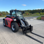 Red Manitou telehandler with long forks attached, parked on a concrete surface with trees and a fence in the background.