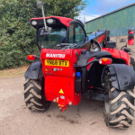 Rear view of a red Manitou telehandler with large off-road tires and tool attachments, parked beside a green corrugated building in a yard.