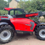Red Manitou telehandler with oversized tires parked outdoors; a person sits in the cab behind the glass.