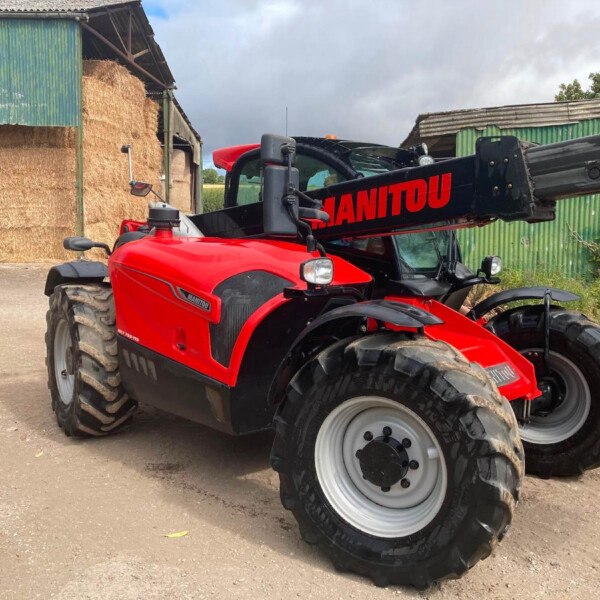 Red Manitou telehandler with extended arm parked near stacked hay bales in a farm shed area under a cloudy sky.