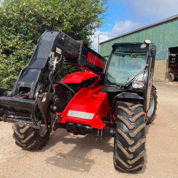 Red Manitou telescopic loader with a large black bucket attached, parked on a dirt lot beside a farm shed with hay bales nearby and leafy trees behind.