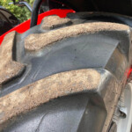 Close-up of a large dirt-covered tire tread with red equipment visible in the background.