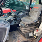 Interior of a Manitou tractor cab with a contoured gray seat and steering wheel; control panel and floor pedals visible, muddy floor mats, and stacked green pipes seen through the window.