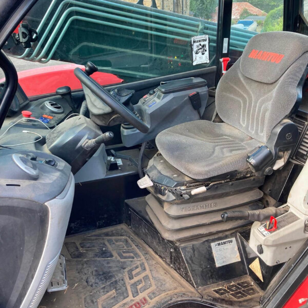 Interior of a Manitou tractor cab with a contoured gray seat and steering wheel; control panel and floor pedals visible, muddy floor mats, and stacked green pipes seen through the window.
