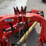 Close-up of red agricultural machinery with hydraulic hoses, metal frame, and warning stickers on a concrete surface.